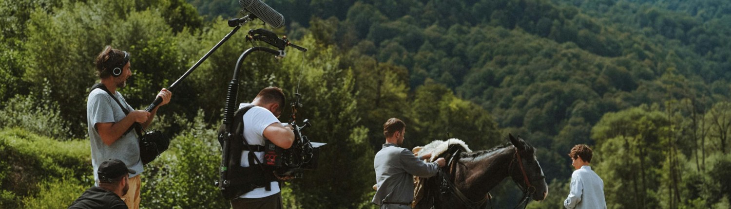 A film crew, actors and a horse on an outdoor set, with a wooded hillside in the background